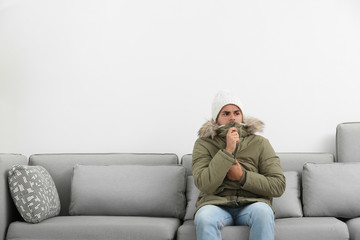 Young man in warm clothes freezing on sofa against white background