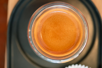 Hot espresso coffee in a clear glass placed on a metal tray and a small wooden shelf on a dark brown wooden table in a vintage style coffee shop.
