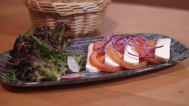 4K Camera pan of a food plate with sliced cheese, tomatos, onions and salat and a bread basket. Standing on a table. Shot with a wide aperture.