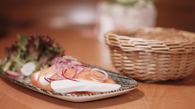 4K shot of a food plate with sliced cheese, tomatos, onions and salat and a bread basket. With a focus pull. Shot with a wide aperture.