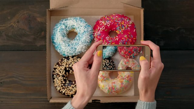 Top view - Woman's hands take pictures with smartphone of delicious fresh donuts in box on wooden table. 4k. Concept of food photo blogging.