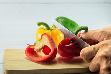 Closeup of woman's hand slicing Red, Yellow, Green, bell pepper or sweet pepper or capsicum on chopping wood on white background with selective focus,raw food ingredient concept.