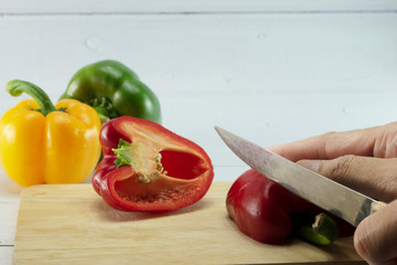 Closeup of woman's hand slicing Red, Yellow, Green, bell pepper or sweet pepper or capsicum on chopping wood on white background with selective focus,raw food ingredient concept.