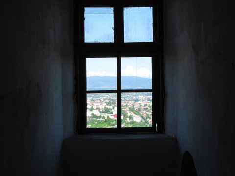 A window from a dark gloomy room into a bright sunny world. Old shabby window with dirty glasses, the dark walls and beautiful cityscape against the backdrop of the mountains outside