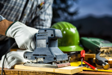 Adult carpenter craftsman wears protective leather gloves, with the electric sander smoothes a...