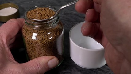 Closeup of a man’s hands transferring a spoonful of instant coffee granules from a glass storage jar into a plain white cup. - Powered by Adobe