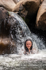 A beautiful smiling girl is standing under the waterfall and the splashes from her head are scattered to the sides. European girl with long black hair. Water falls from large granite stones. Vertical.