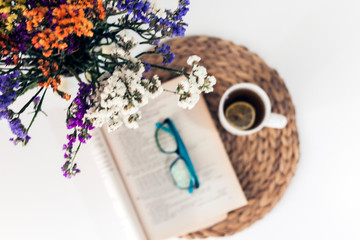 bouquet of flowers, open book and glasses, lemon tea in a white ceramic cup on a wicker stand on the table, top view.