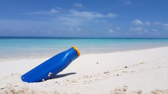 A sun screen bottle rests on white sand on a tropical beach with small waves washing onshore in Hawaii