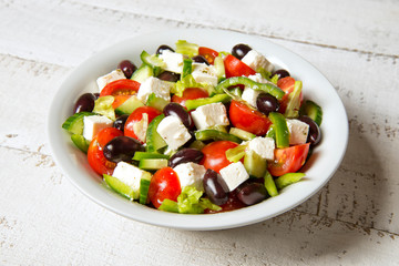 Traditional Greek salad in a round white salad bowl on an white wooden table. Selected focus.