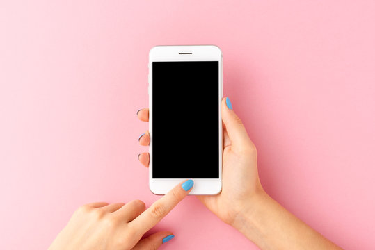 Overhead shot of woman’s hands holding mobile phone with empty screen on pink background. Top view