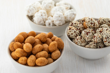 Peeled peanuts, peanuts in syrup sprinkled with sesame seeds and peanuts in sugar in three white bowls on a white wooden background, side view from above