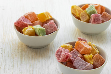 Three white glass bowls with multi-colored pieces of Turkish delight on a white wooden surface, side view from above