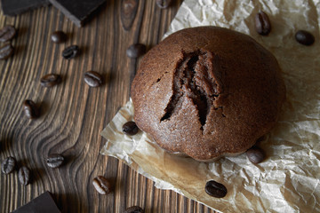 One homemade chocolate muffin on parchment with coffee beans and pieces of chocolate. Dark wooden background, closeup, top view
