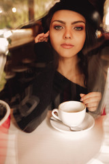 beautiful young curly woman in hat with cup of coffee in cafe through glass