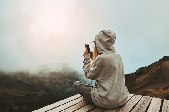a girl in a grey hoodie sitting on a viewpoint and take a picture of sunset on smart phone
