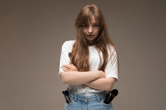 A young beautiful girl with long wavy rustic hair, nud makeup, in a white jersey, stands crossing her hands, and there are lots of makeup brushes in her pockets gin