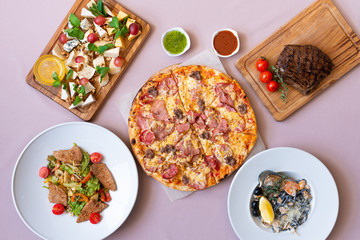 Dining table with food, top view. Pizza, salad, cheese board, steak, black seafood pasta.