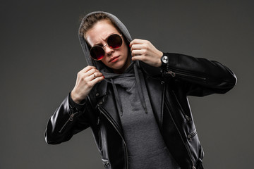 Obraz premium A young girl with rough facial features, patted brown hair, bright manicure, in a grey bike, black jacket, holds sunglasses with her hands, puts a hood on her head and holds it with her hands