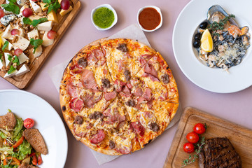 Dining table with food, top view. Pizza, salad, cheese board, steak, black seafood pasta.