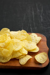 Crispy potato chips with salt on a rustic wooden board on a black surface, side view. Space for text.