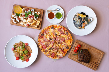 Dining table with food, top view. Pizza, salad, cheese board, steak, black seafood pasta.
