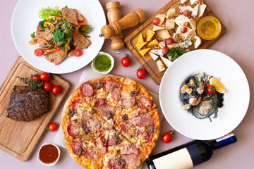 Dining table with food, top view. Pizza, salad, cheese board, steak, black seafood pasta, bottle of wine.