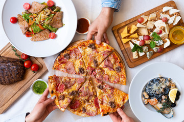 The company of friends takes slices of pizza on the table with a varied meal, top view. Eating and leisure concept.