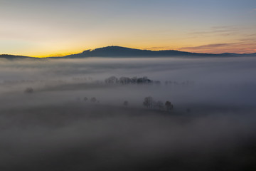 Fototapeta na wymiar Aerial view to autumn foliage trees silhouette in misty fog and hill in sunrise, Czech landscape