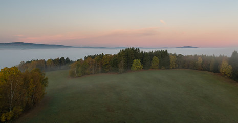 Aerial view to autumn foliage trees and meadow in misty fog and hill at sunrise, Czech landscape