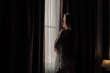 A beautiful girl looks out the window covering her body with a man's shirt in the dark