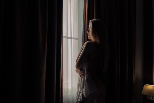 A beautiful girl looks out the window covering her body with a man's shirt in the dark