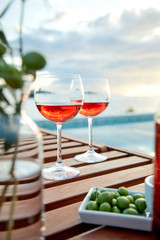 two glasses of rose wine with  olives against blue water on a pier..