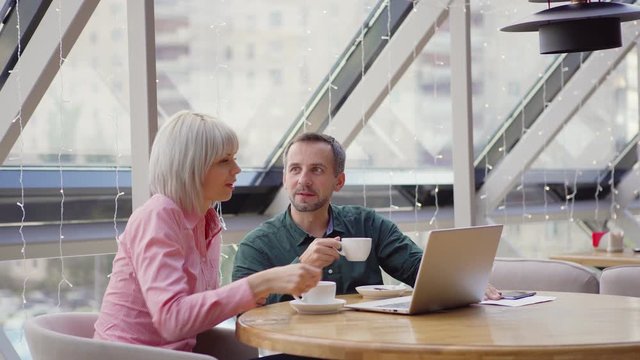 Man and woman in casual outfits sipping hot drinks while discussing data from laptop sitting at restaurant table and collaborating on new project together