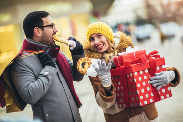 Young couple dressed in winter clothing holding gift boxes outdoor. They eat pretzels.