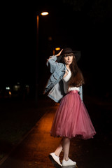 Young woman in black hat, jeans jacket, white tank top, pink chiffon skirt and white sneakers on a street at night