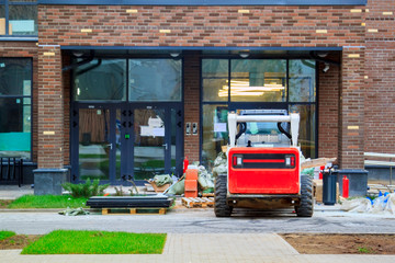 Fototapeta premium Skid loader stands in front of entrance to new multi-storey house and waits for garbage to be loaded into bucket. Mini bulldozer waiting for cargo in front of porch
