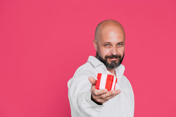 Handsome serious middle-aged man in a white shirt holds a red gift in his hand decorated with a white bow smiling on a pink background with copy space