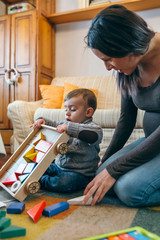 Young mother playing with her toddler son a wooden game building in the living room