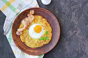 Potato hash brown with egg and bacon on a brown clay plate on a dark concrete background. American cuisine. Top view.