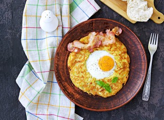 Potato hash brown with egg and bacon on a brown clay plate on a dark concrete background. American cuisine. Top view.