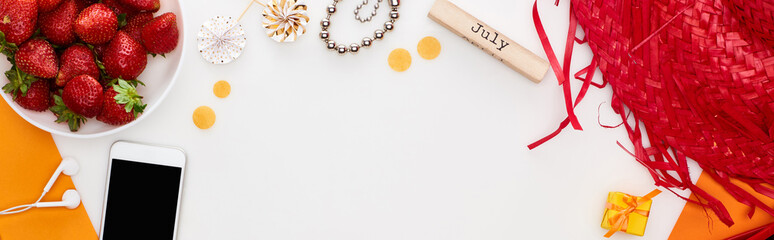 panoramic shot of wooden block with july inscription, smartphone, fresh strawberry, straw hat, lollipops, earphones, bracelet isolated on white