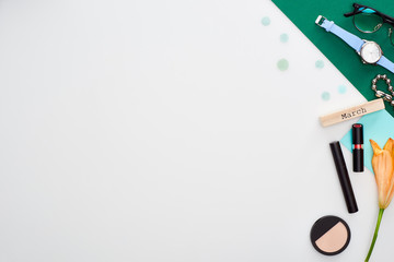 lipstick, mascara, face powder, wooden block with march inscription, glasses, wristwatch, lily flower, green paper isolated on white