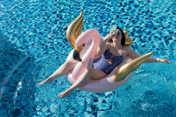 Attractive young woman takin a selfie sitting on the pink flamingo / swan pool float in the swimming pool with