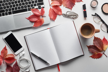 top view of laptop near smartphone, coffee cup, cosmetics, earphones, glasses, notebook and red leaves of wild grapes on white table