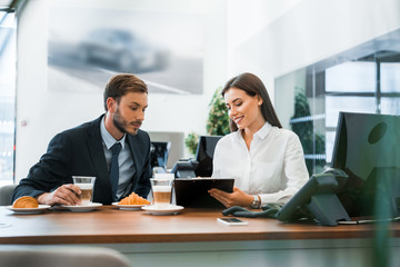 selective focus of happy car dealer holding clipboard near man and tasty croissants