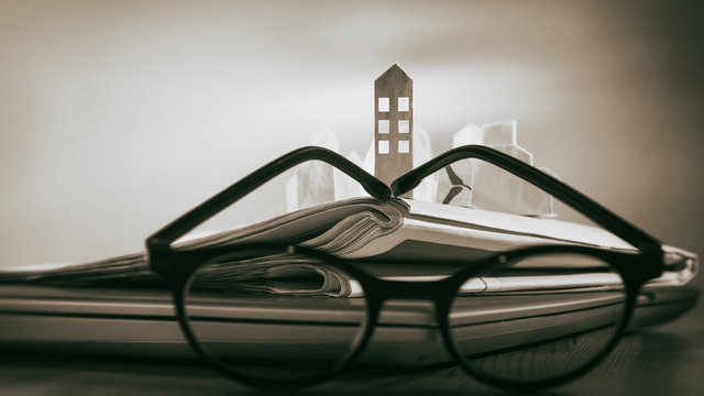 Magnifying glass in front of an open newspaper with paper houses. Concept of rent, search, purchase real estate.