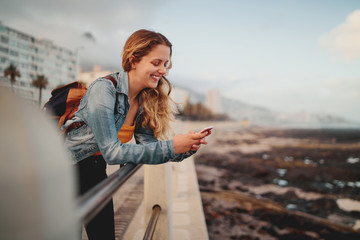 A smiling blonde young female traveler with her backpack leaning on railing at city street using...