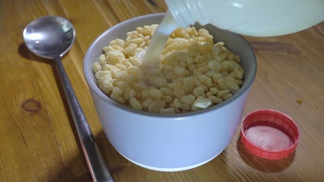 Normal speed then slow motion: Close POV shot of skimmed milk being poured from a plastic bottle onto dry crisped rice breakfast cereal in a bowl, with a spoon alongside.