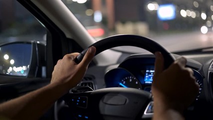Man driving car in a night city. Male hands holding steering wheel. Oncoming traffic is out of focus with beautiful bokeh. 4K - Powered by Adobe